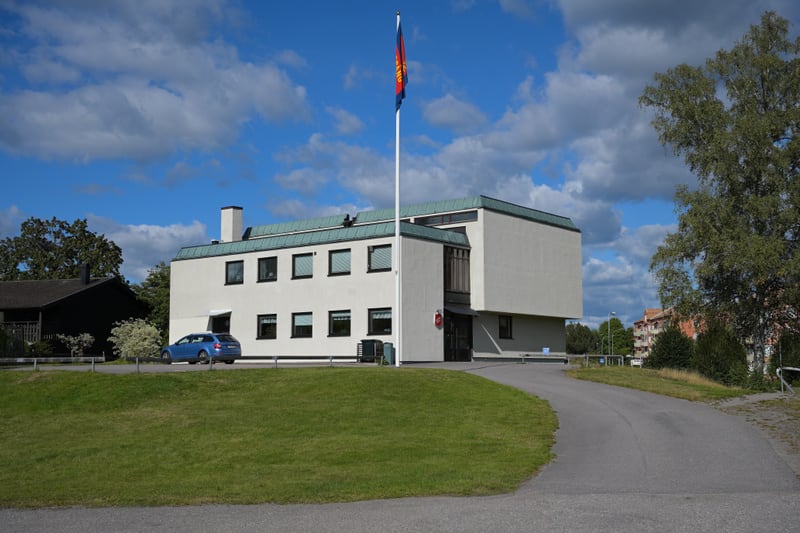 En modern byggnad med ljus fasad och mörkare tak dominerar bilden under en blå himmel med moln. En flaggstång med flagga står framför byggnaden och en blå bil är parkerad. Grönt gräs och träd omger byggnaden, samt en asfalterad väg.
