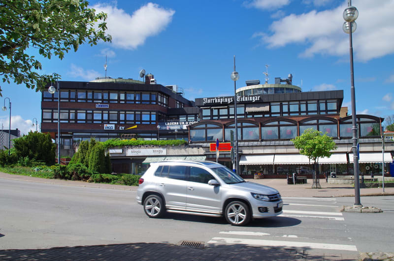 En silverfärgad SUV kör på en asfalterad väg framför en större byggnad med skylten "Norrköpings Tidningar". Byggnaden har flera fönster och på nedervåningen finns butiker med markiser utrullade. Ett träd med gröna löv syns i förgrunden och himlen är