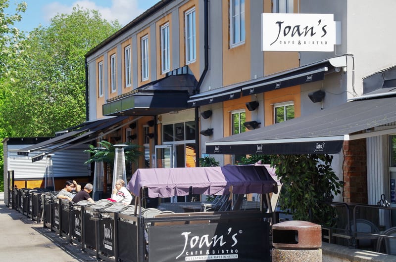 En bild som visar byggnaden som huserar "Joan's Cafe & Bistro" med dess uteservering. Flera personer syns sitta vid borden på uteserveringen, som är omgiven av svarta skärmar. Byggnaden har fönster och svarta markiser.