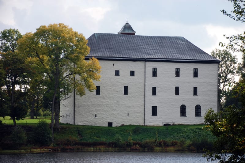 En stor vit byggnad med ett mörkt tak och ett mindre torn med kors på sitter upphöjd över en vattenmassa. Träd med löv i gröna och gula nyanser omger byggnaden, vilket tyder på övergången från sommar till höst.