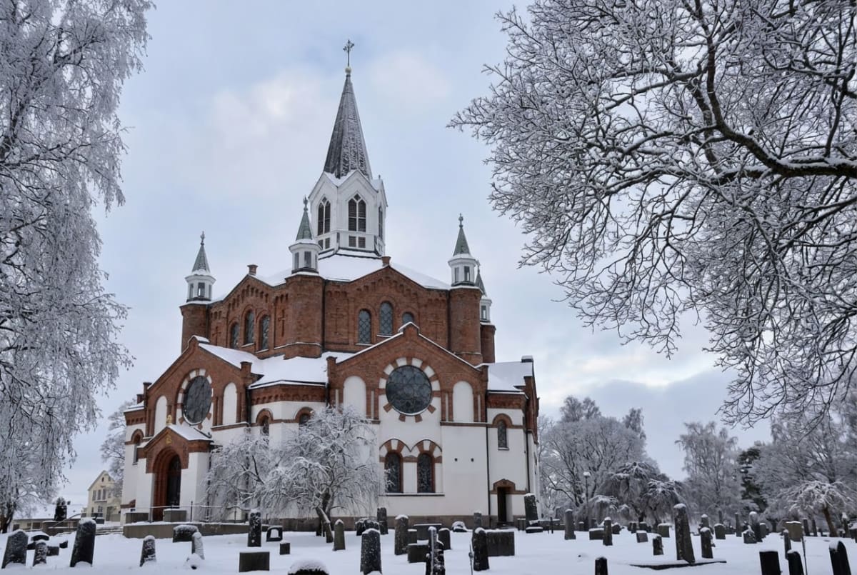 En stor röd kyrka med vitt tak och snö täcker marken och grenarna på träden. På kyrkogården runt kyrkan står flera gravstenar som är delvis täckta av snö under en gråmulig himmel.