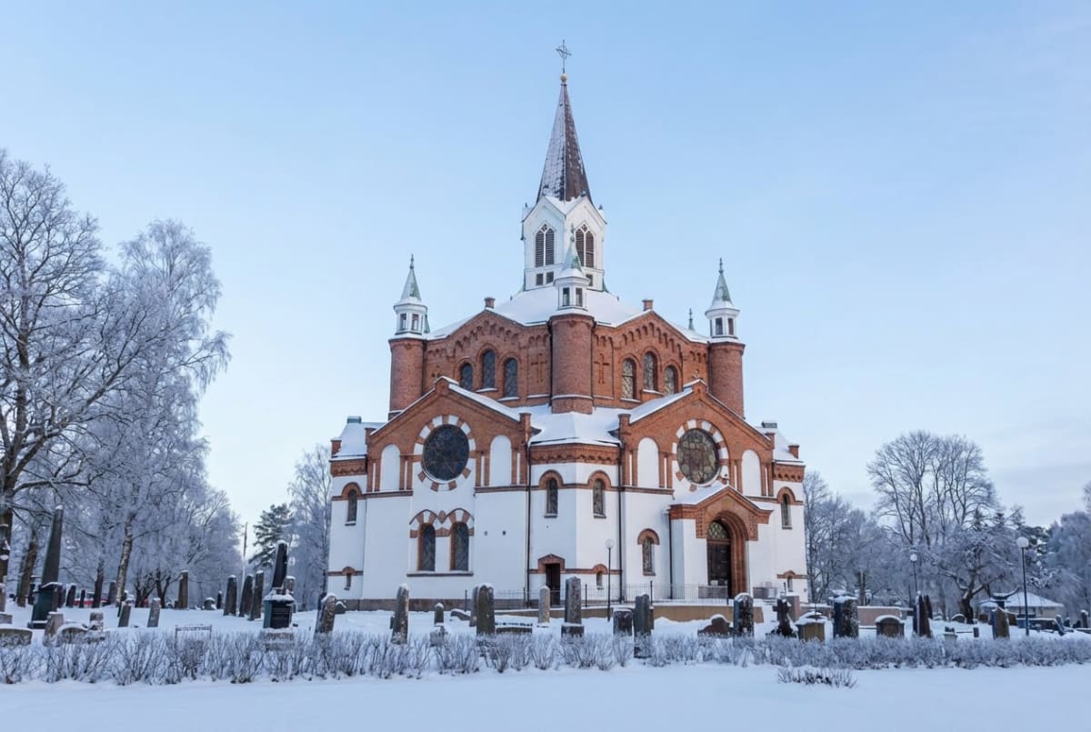 En stor kyrka med röd tegelfasad och vita partier, täckt med snö på taket och marken. Runtomkring finns en kyrkogård med gravstenar och snötäckta träd med rimfrost. Himlen är klarblå.