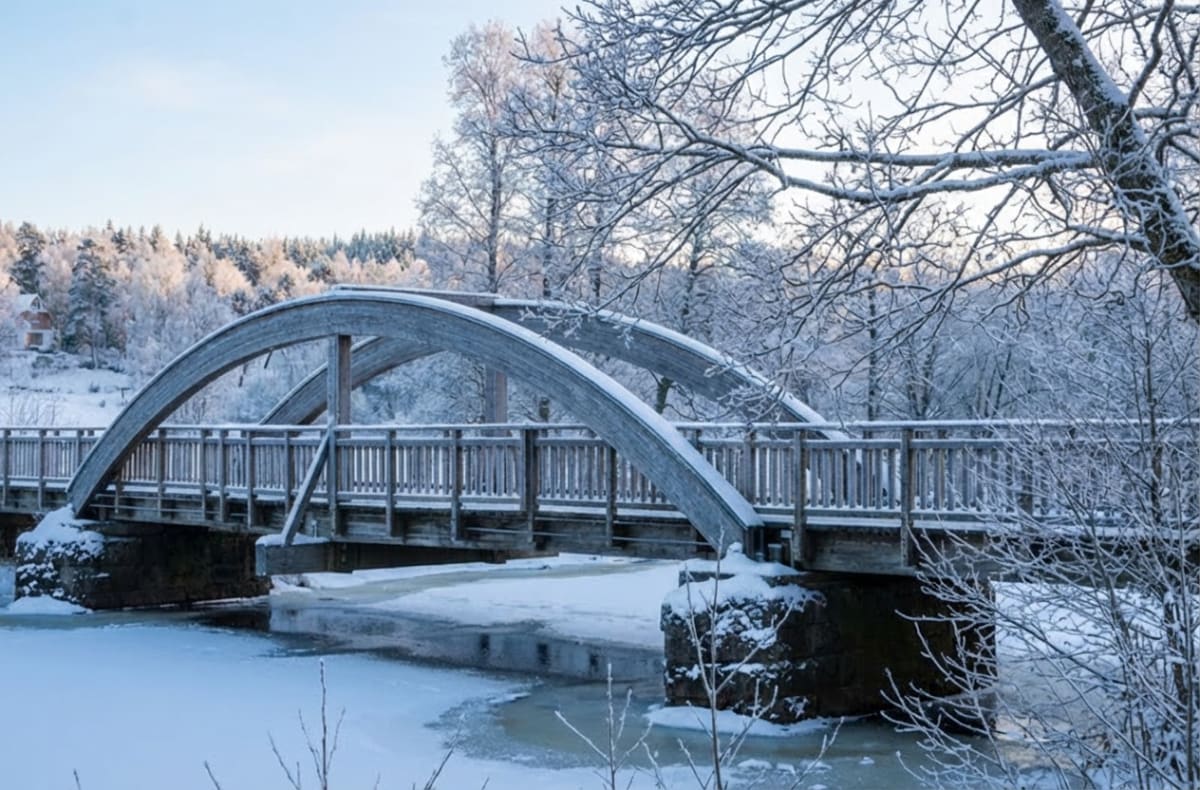 En vinterbild föreställande en träbro med två valv över en delvis frusen å. Naturen runtomkring är täckt av snö och frost, med snötäckta träd i bakgrunden och en liten byggnad synlig på en kulle till vänster.