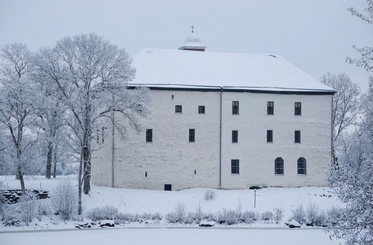 En stor vit byggnad med ett snötäckt tak och en liten tornhuv med ett kors ses i bildens mitt. Framför byggnaden finns snötäckta träd och buskar, och i förgrunden syns en frusen yta, troligen is eller snö. Höga träd täckta av snö står på vänster sida