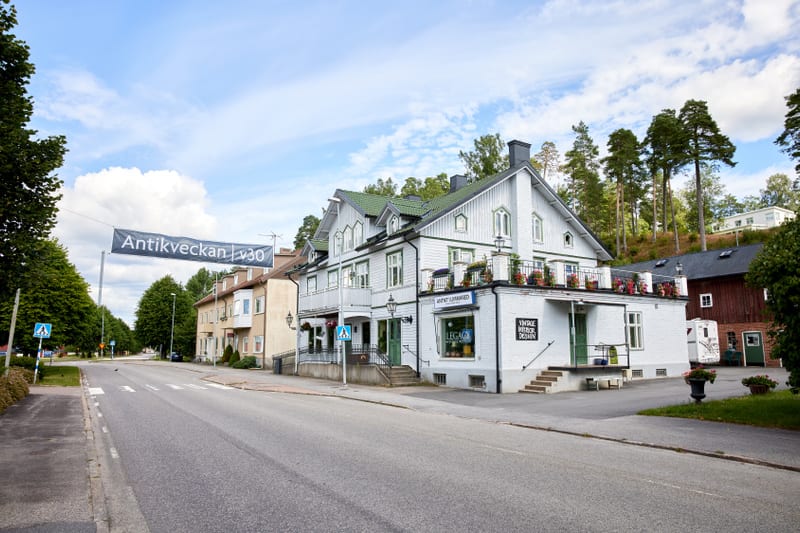 En gata med parkerade bilar och hus på båda sidor. Ett vitt hus med grått tak står i förgrunden till höger, med skyltar med texten "Antioch Learner" och "Vintage Interior Design". En stor banderoll med texten "Antikveckan v30" hänger över gatan. En ö