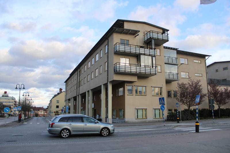 En gata med ett stort beige färgat hus med balkonger i förgrunden till höger. En silverfärgad kombibil kör på gatan. I bakgrunden syns fler byggnader och en blå himmel med vita moln.