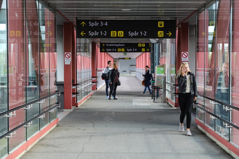 En bild som visar en modern gångbro, sannolikt vid en tågstation eller bussterminal, upplyst av naturligt ljus. Personer syns gående och ståendes med skyltar som hänvisar till "Spår 3-4" och "Spår 1-2" samt "Ersättningsbuss". En skylt visar en symbol