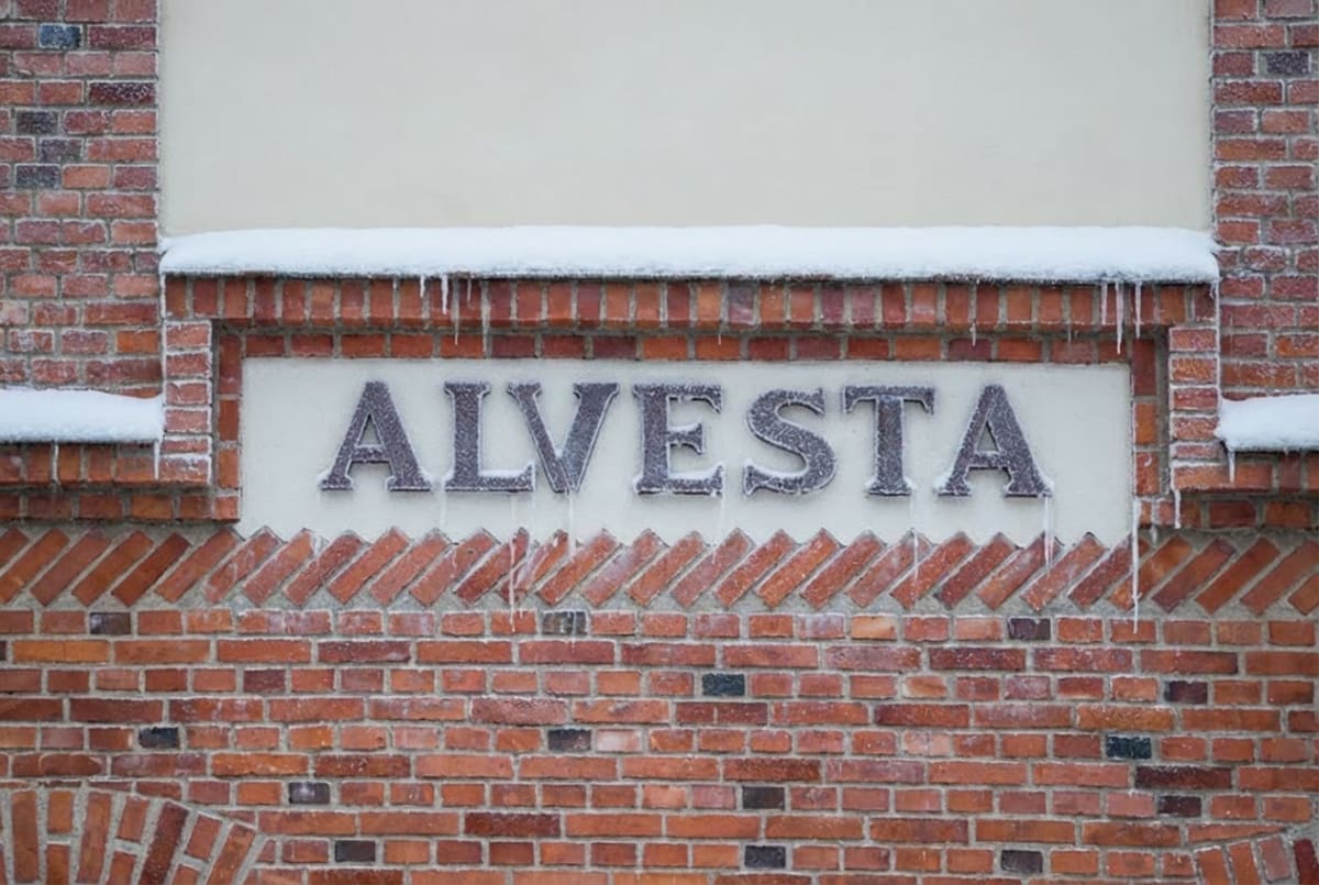 En närbild av en tegelfasad med vita detaljer. Centrum av bilden pryds av texten "ALVESTA" i stora, svarta bokstäver som är täckta av snö och frost. Ovanför och under texten finns dekorativa tegelsättningar och snö samt istappar hänger från kanten ov