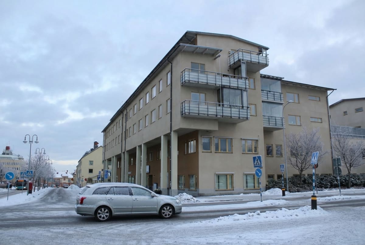 En vinterbild tagen utomhus under dagtid, som visar en silverfärgad bil som kör på en snötäckt gata framför en större gulbeige byggnad med balkonger. Träd med rimfrost syns längs gatan och himlen är molnig.