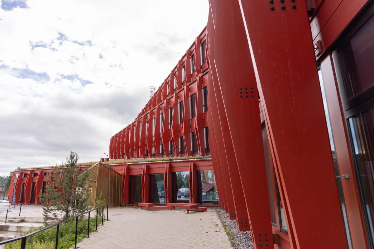 En modern byggnad med röd metallfasad och stora fönster under en molnig himmel. Byggnaden har ett grönt tak och verkar ha flera våningar. Vid byggnaden finns en gångväg, en liten gran och en metallräcke.