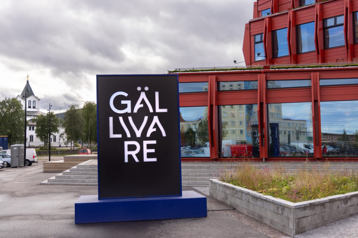 En stor svart skylt med vit text "GÄLLIVARE" står i förgrunden, med en röd byggnad med stora fönster i bakgrunden. Till vänster syns delar av en vit byggnad med ett torn och kors. Marken är grå och himlen är molnig.