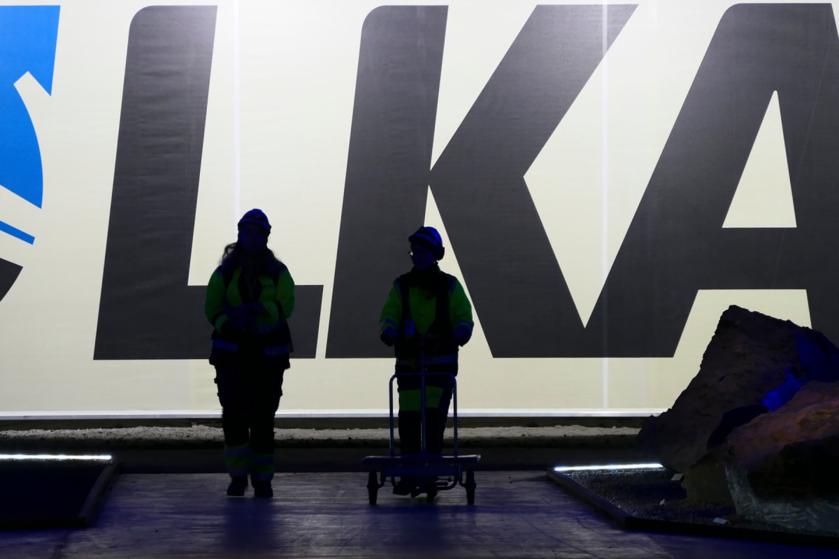 Två personer i profil, klädda i skyddskläder, skymtar i förgrunden. De står framför en stor vit bakgrund med stora svarta bokstäver som bildar ordet "LKAB". En av personerna står vid en vagn. Belysning underifrån ger en silhuett-effekt och skapar mör