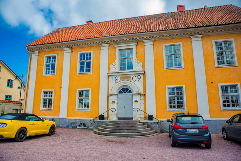 En gul byggnad med rött tak och vita detaljer står under en blå himmel med moln. Framför byggnaden finns en uppfart täckt med grus. Två bilar är parkerade, en gul cabriolet till vänster och en mörkgrå kombi till höger. På byggnaden syns årtalet '1809