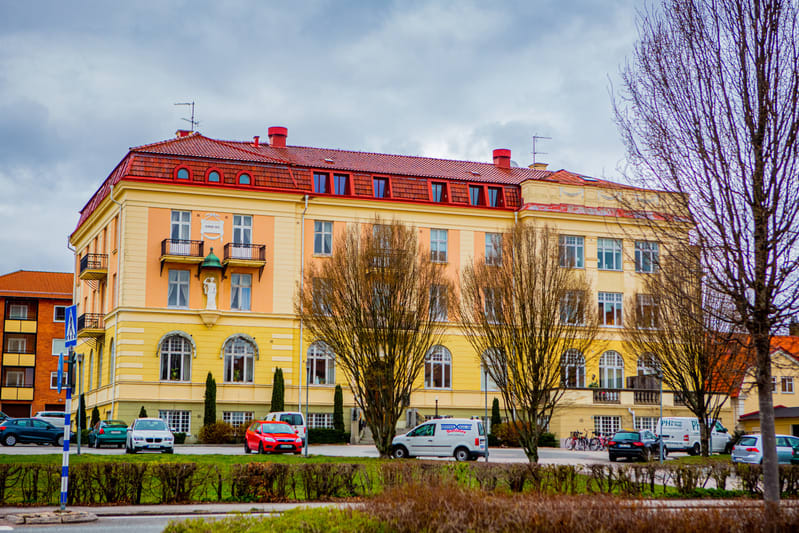 En gul byggnad med rött tak och prydnadsbalkonger står under en molnig himmel. Framför byggnaden syns kala träd, en gräsyta med häckar, och parkerade bilar i olika färger. En blåvit skylt sticker upp på vänster sida.