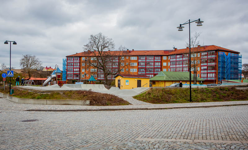 En bild som visar en miljö utomhus med en stor tegelbyggnad med balkonger i rött, en skateboardpark, lekplats och flera träd. Förgrunden består av en stenlagd gata. Himlen är grå och mulen. Delar av byggnaden är täckt med byggnadsställningar och blåa