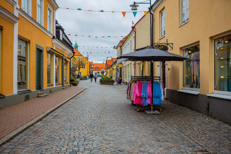 En kullerstensgata kantad av butiker i färgglada hus under en gråmulen himmel. En klädställning med färgrika kläder och ett parasoll står intill en butik. I bakgrunden syns människor och en kyrktorn. Flaggspel hänger över gatan.