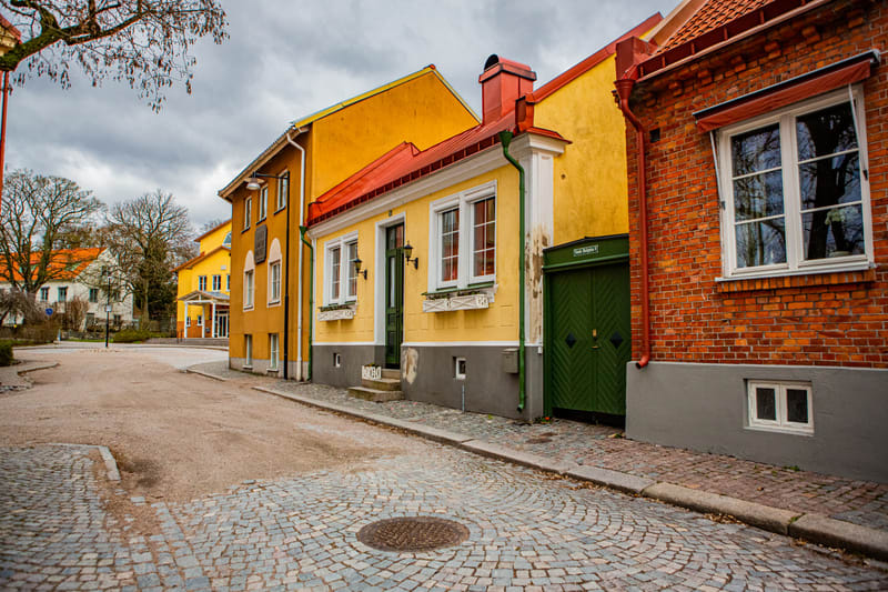 Bilden visar en gata med kullersten i förgrunden och pastellfärgade hus med röda tak under en molnig himmel. Två gula hus syns tydligt, varav ett med en grön dörr och vita fönster. Till höger syns en tegelbyggnad med vita fönster. Ett smalt träd med