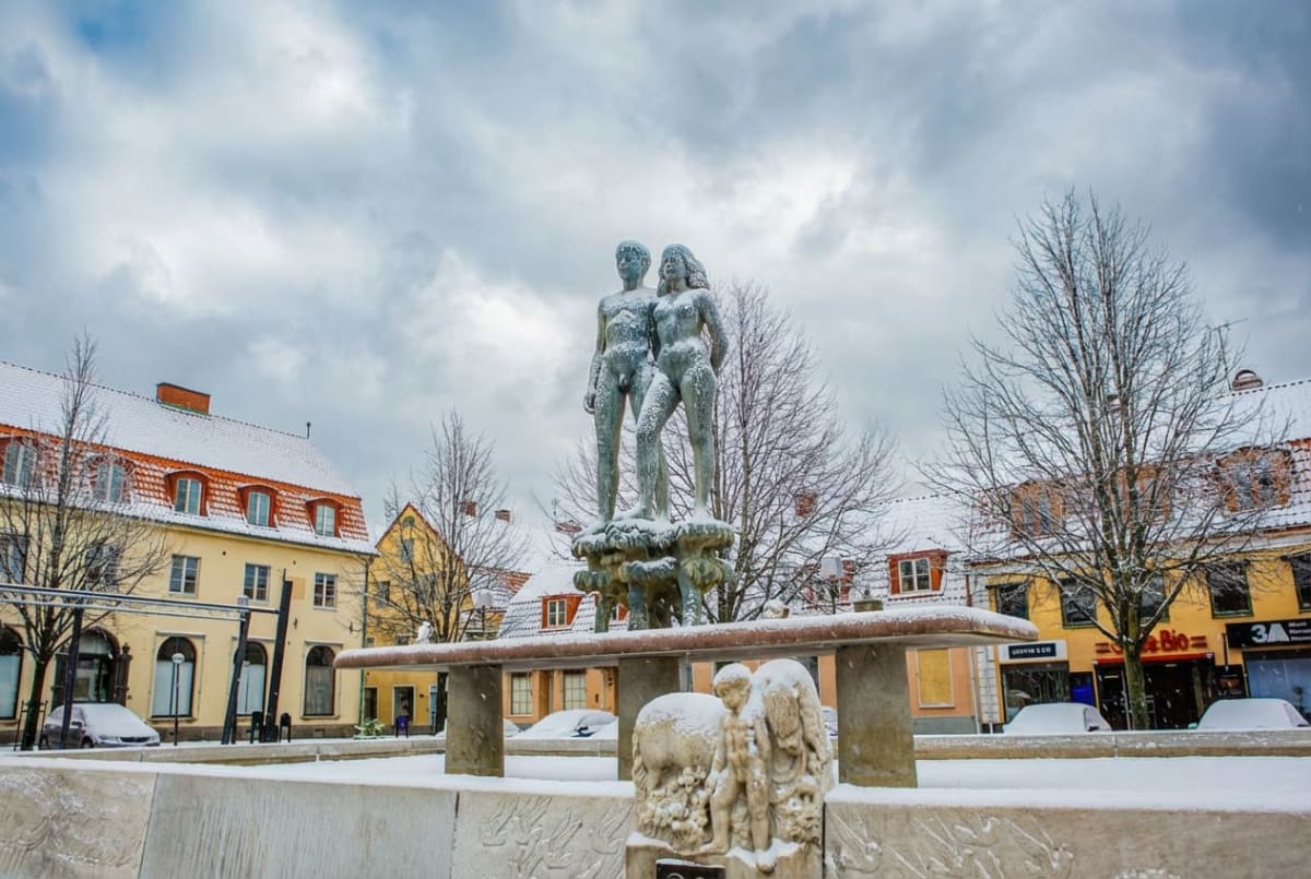 En ganska bred bild av ett torg täckt med snö där en staty med två människor i relief i mitten är fokus. Hus med snötäckta tak och kala träd syns i bakgrunden, samt parkerade bilar delvis täckta av snö.
