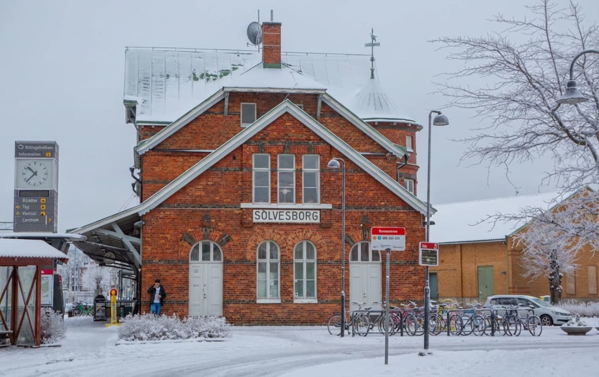 En röd tegelbyggnad, med texten "SÖLVESBORG" ovanför tre stora fönster, står omgiven av nysnötäckt mark under en grå vinterhimmel. Taket på byggnaden är också snötäckt. Framför byggnaden finns cykelställ med flera cyklar, en klocka med skyltar och en