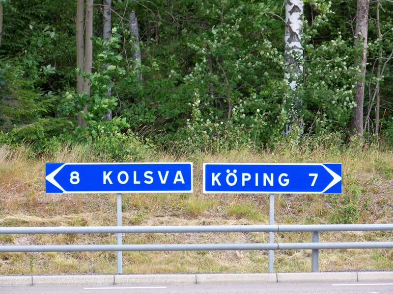 Två blå vägskyltar syns intill en vägkant. Den vänstra skylten pekar mot Kolsva, 8 km bort, och den högra mot Köping, 7 km bort. I bakgrunden syns skog och gräs. En grå vägräcke löper framför skyltarna och en bit av vägbanan syns i förgrunden.