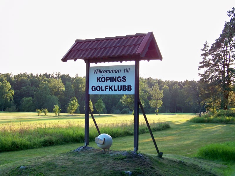 En utomhusbild föreställande en skylt med texten "Välkommen till KÖPINGS GOLFKLUBB" framför ett grönskande landskap med träd och en klippt gräsmatta. En golfbolls-liknande figur med golfklubba syns vid skyltens fot.