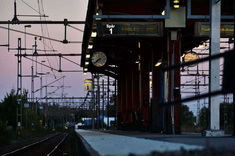 En bild av en tågperrong i skymning eller gryning. Perrongen har ett tak med belysning och två klockor, samt en digital skylt som visar "Spår 1". Till vänster om perrongen syns järnvägsspår och kontaktledningar. Himlen är lila och rosa.