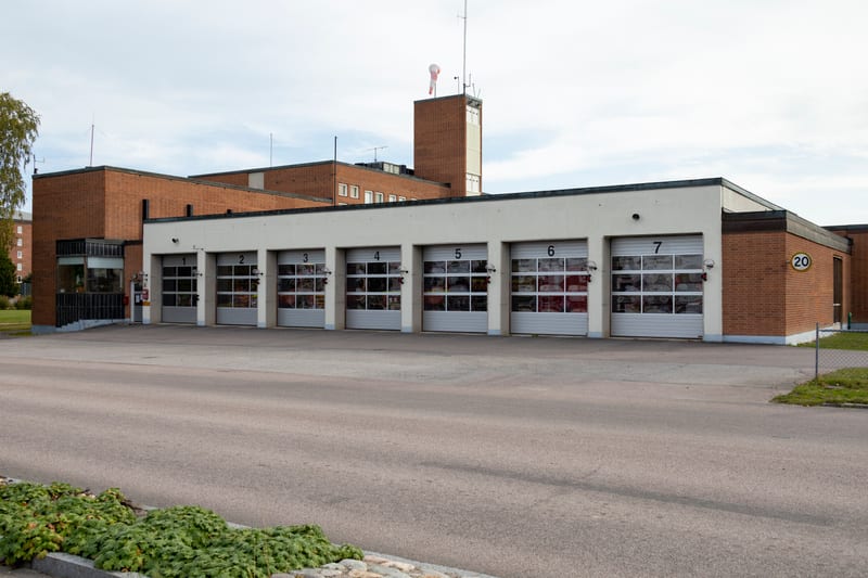 En stor röd tegelbyggnad med en lägre vit tillbyggnad som har sju garageportar. På taket syns en vindstrut. Marken framför byggnaden är asfalterad, och i förgrunden finns en gräsmatta med vegetation och stenar. Ett träd syns till vänster i bilden.