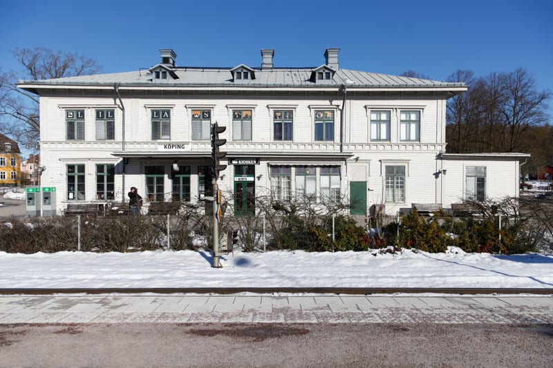 En ljus byggnad med flera fönster och dörrar står under en klarblå himmel. Framför byggnaden finns snötäckta markpartier, buskar och en asfalterad yta med en skymt av räls. Skylten "KÖPING" syns på byggnaden.