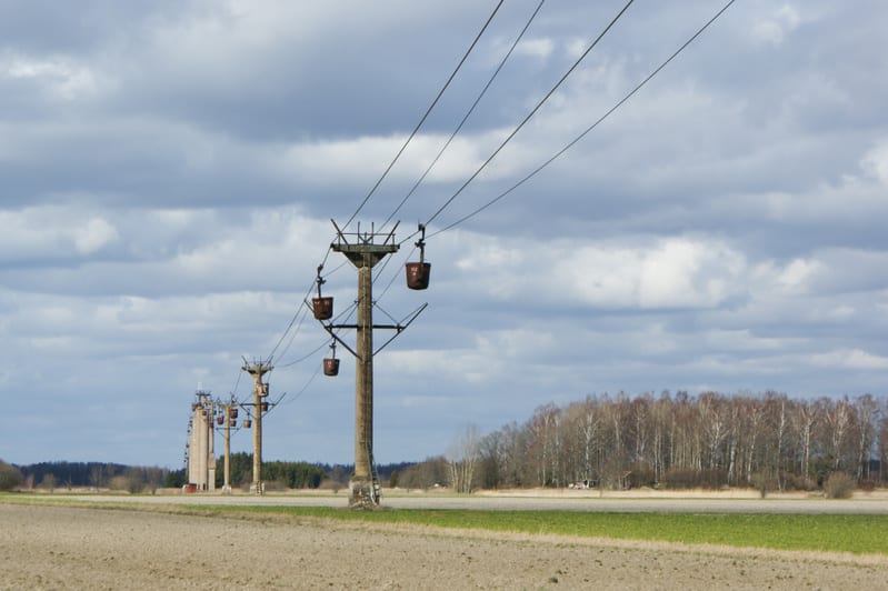 En rad stående stolpar till en linbana med hängande korgar sträcker sig över ett öppet fält under en molnig himmel. I bakgrunden syns skog med kala träd.