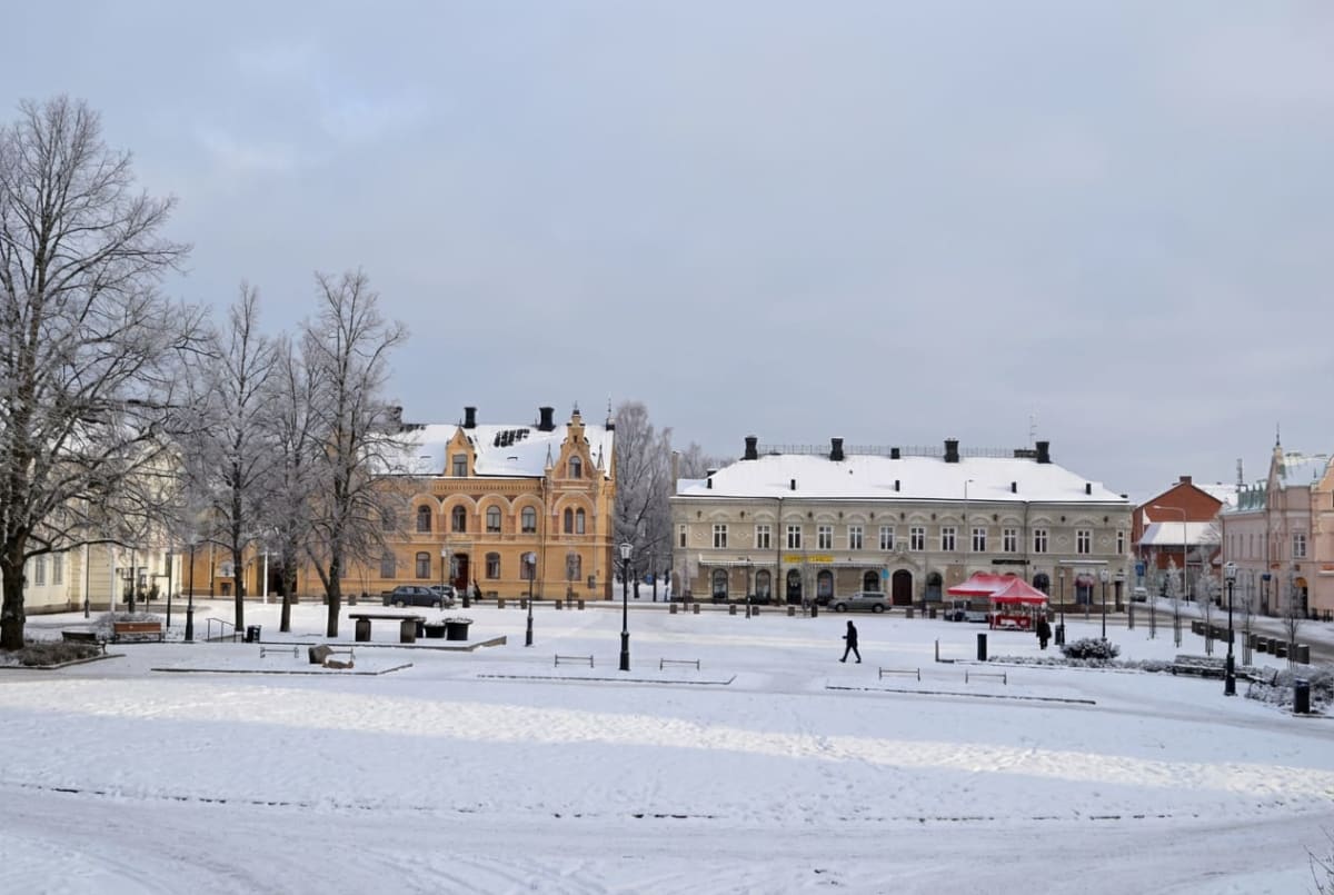 En vinterbild tagen utomhus vid dagtid, som visar ett snötäckt torg med byggnader och träd. En person promenerar över torget, och några fordon är parkerade i bakgrunden. Träden är kala och täckta med frost, och taken på byggnaderna är snötäckta.
