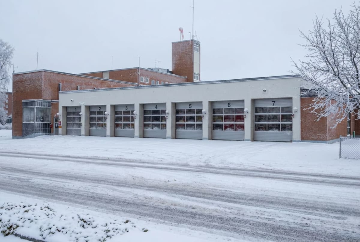 En röd tegelbyggnad delvis klädd i vit fasad med sju nummererade garageportar syns i ett snöigt vinterlandskap. En skorsten med väderskärm och antenner står på byggnadens tak. Framför garagen finns en snötäckt uppfart med däckspår.