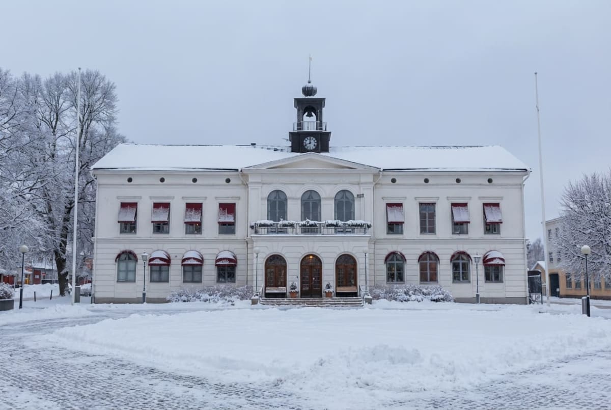 En stor vit byggnad framifrån under vintertid. Byggnaden har två våningar med runda och rektangulära fönster, prydda med röda markiser. Mitt på taket står ett klocktorn, och framför byggnaden ligger ett snötäckt område med spår. Träd med snötäckta gr