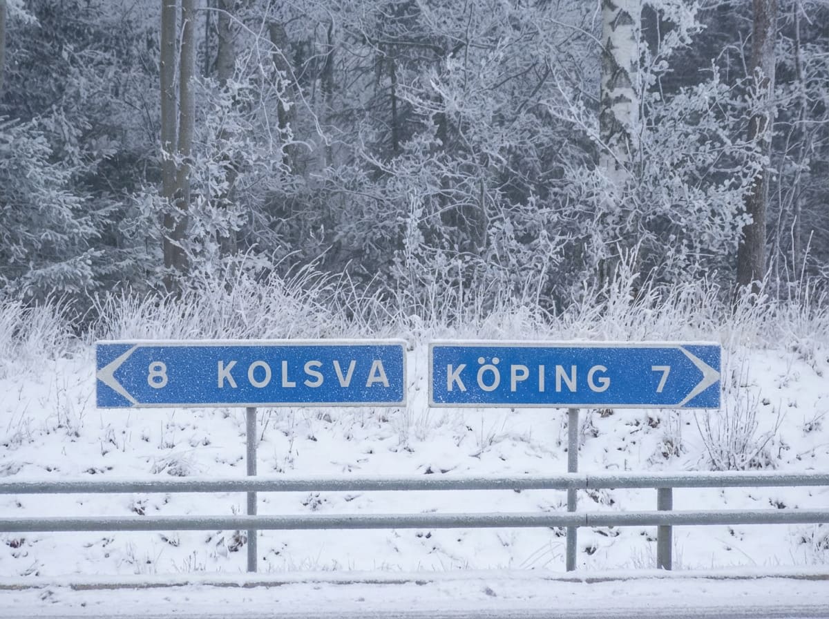 Två blå vägskyltar syns på en vinterväg. Skyltarna, 'KOLSVA 8' och 'KÖPING 7', är täckta av snö. Ett snötäckt skogslandskap utgör bakgrunden, och ett snötäckt vägräcke syns framför skyltarna.