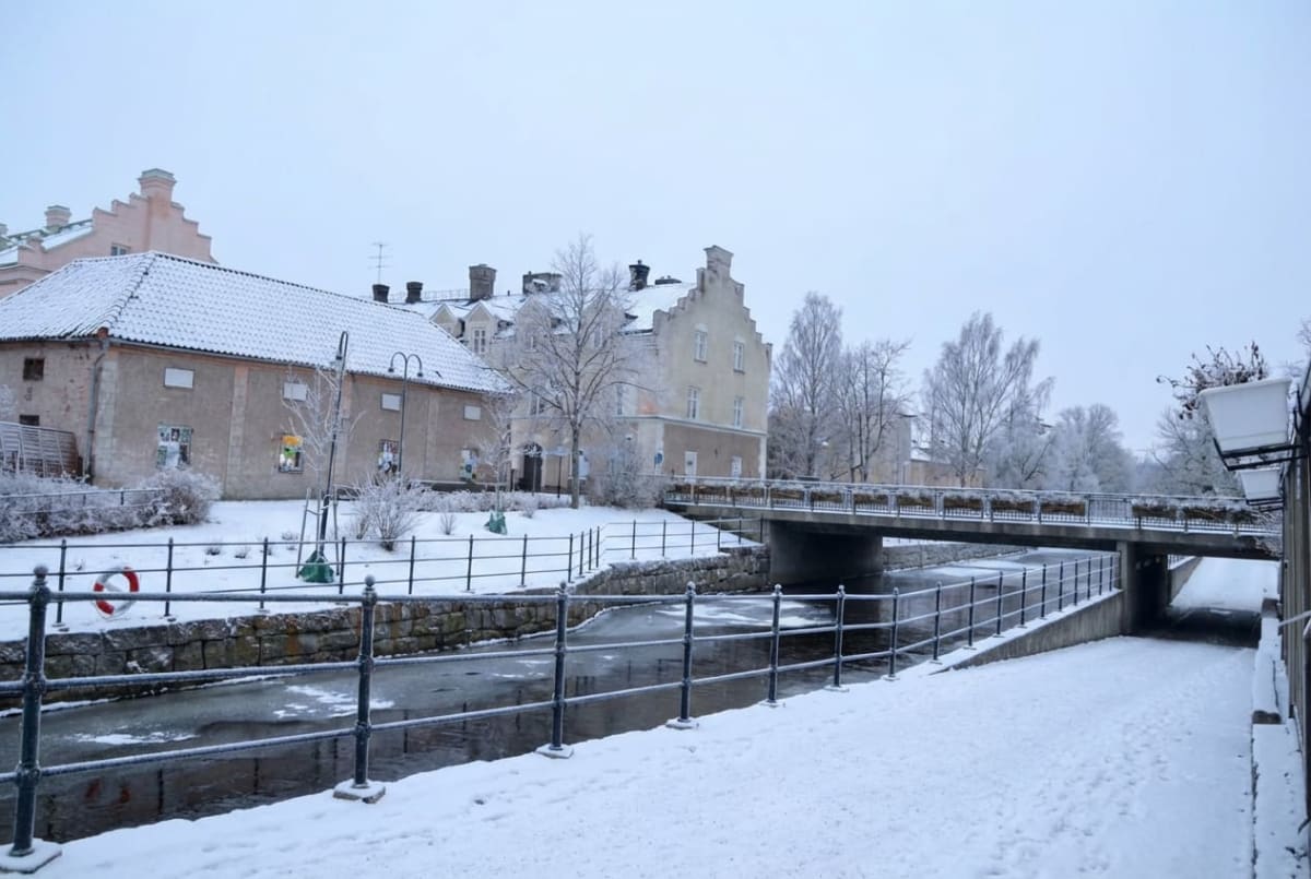 En vinterbild som visar snöklädda markytor, en delvis isbelagd kanal eller å, och en bro i centrum av bilden. Längs vänster sida av vattendraget står byggnader med snö på taken och träden är frostiga. Ett räcke löper längs vattnet.