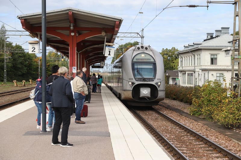 En bild tagen i ögonhöjd som visar en tågstation utomhus, med ett tåg som står vid perrongen medan passagerare väntar. I bakgrunden syns skog och en vit byggnad. En skylt med "Spår 1" syns vid perrongen och en annan med "Spår 2" i bakgrunden.