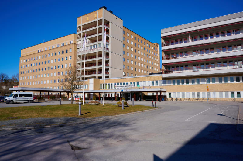 En stor sjukhusbyggnad i flera våningar med gulbeige fasad och många fönster syns under en klarblå himmel. Framför byggnaden finns en asfalterad yta, gräsmattor, träd, några bilar och en tydlig skylt med texten ”Västmanlands Sjukhus Köping”.