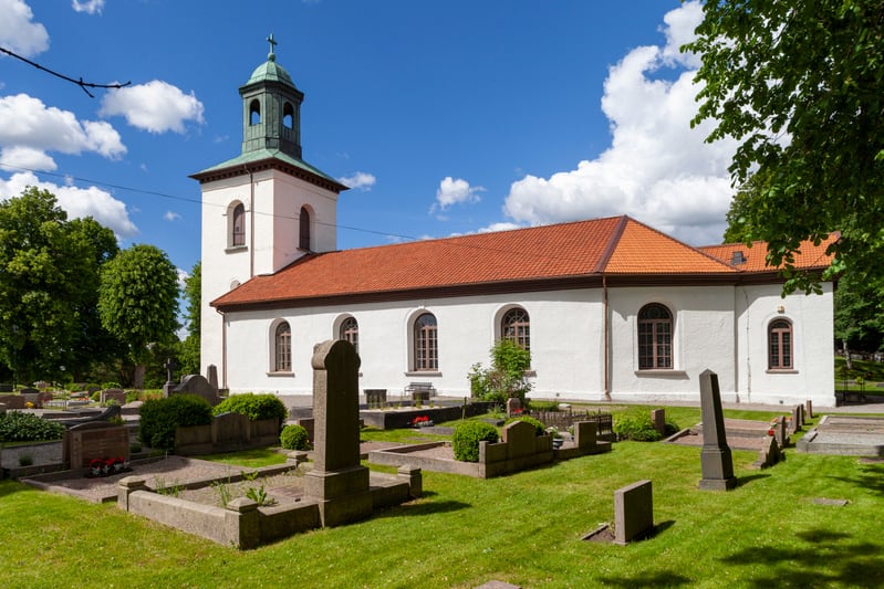 En vit kyrka med rött tegeltak och ett grönt klocktorn syns under en molnig blå himmel. Framför kyrkan breder en kyrkogård ut sig med gräsmattor, stenar och enstaka buskar. Gröna träd ramar in kyrkogården i bakgrunden.