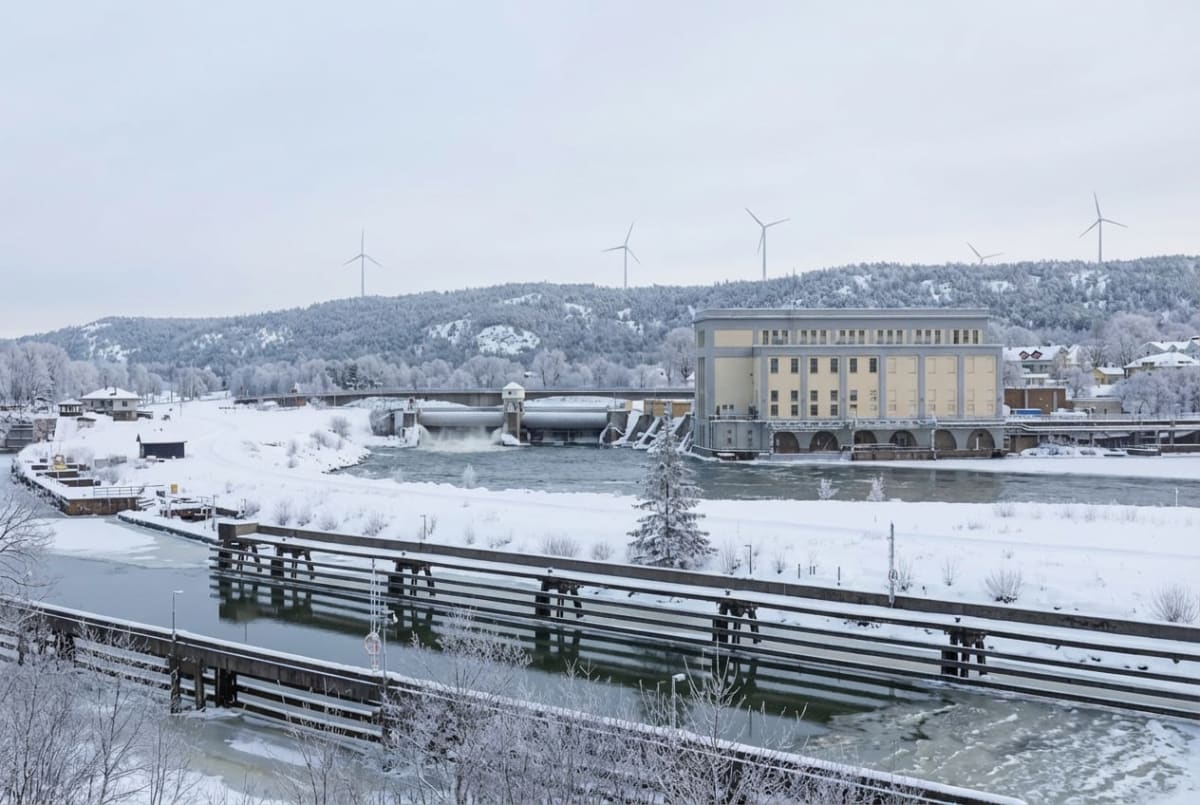 En vintervy över ett vattenkraftverk med en flod och en sluss. Landskapet är täckt av snö och frost. På en kulle i bakgrunden syns flera vindkraftverk, och hus bland de snöklädda träden. En bro korsar floden framför kraftverket.
