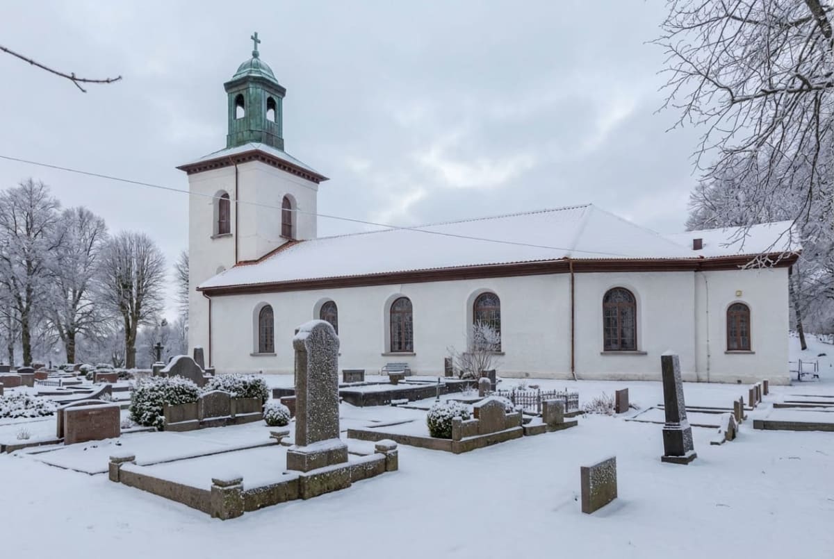 En vit kyrkobyggnad med ett grönt kyrktorn under ett snötäckt tak, omgiven av en snötäckt kyrkogård med gravstenar och träd, under en molnig himmel.