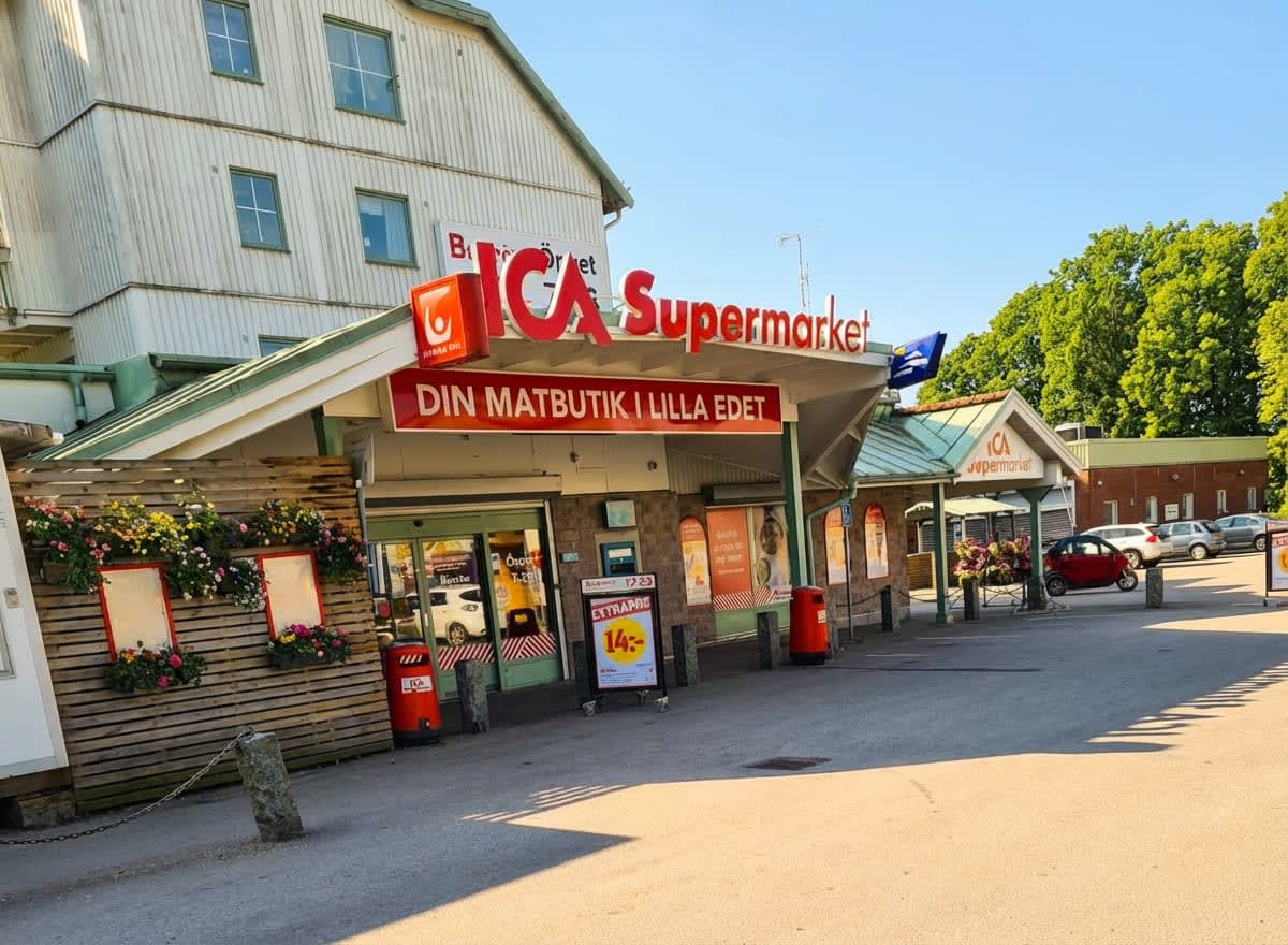 En fasadvy av en ICA Supermarket-butik under dagtid med en skylt som lyder "DIN MATBUTIK I LILLA EDET". Utanför butiken finns parkeringsplatser och träd i bakgrunden. Ett hus med vit träpanel syns ovanför butiken.