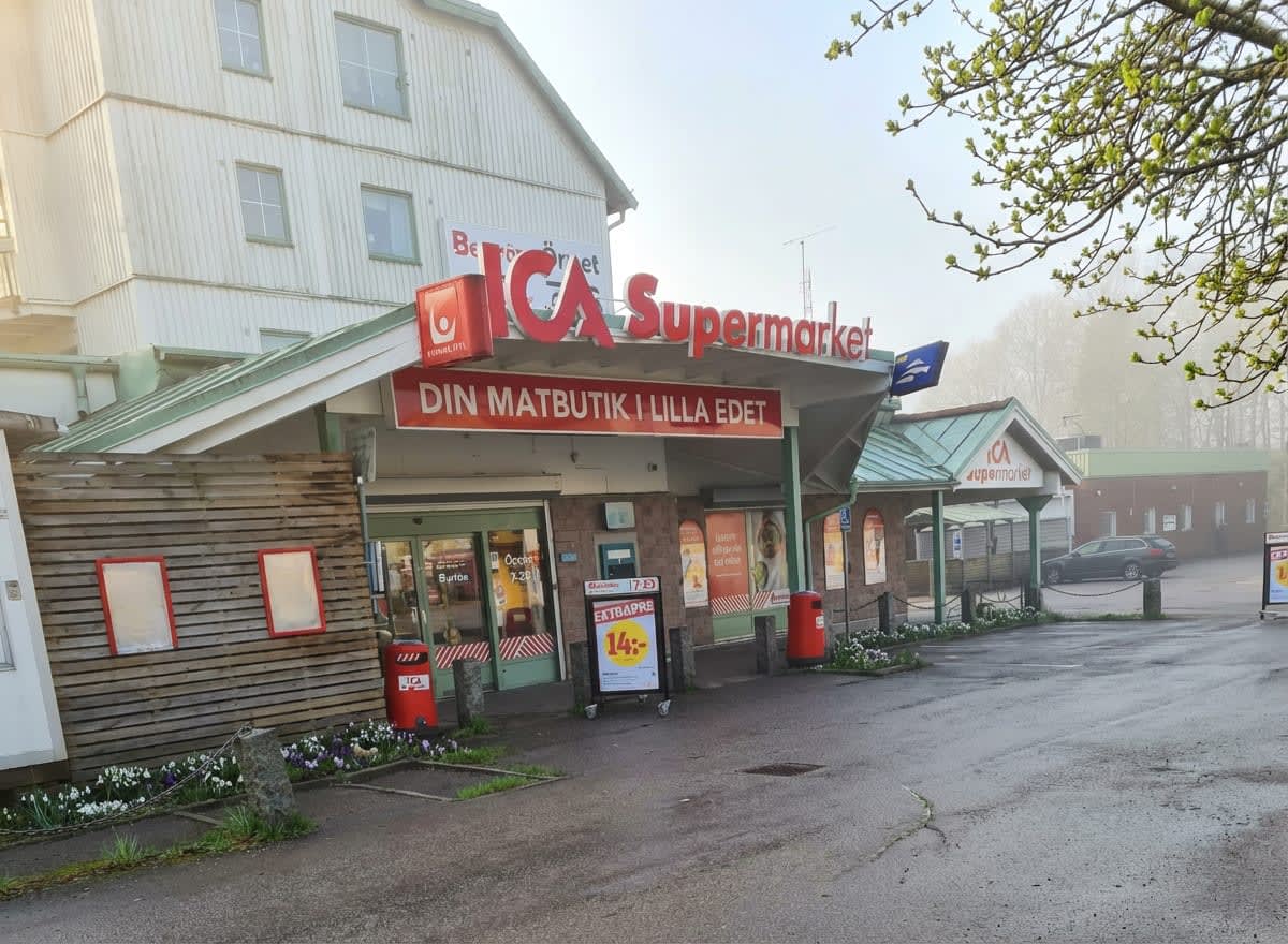 En bild utomhus som visar entrén till en ICA Supermarket-butik med texten "DIN MATBUTIK I LILLA EDET" på skylten. Framför butiken finns en asfalterad yta och nyutslagna knoppar på trädgrenar syns i förgrunden.