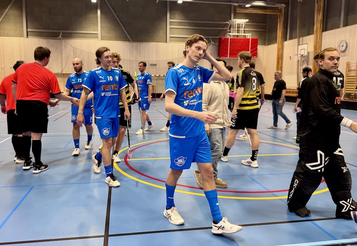 En grupp män i sportkläder befinner sig i en idrottshall, troligen för innebandy. Flera av dem bär blåa matchtröjor med siffror och sponsortryck. En man i röd tröja skakar hand med en annan man. Golvet är blått med linjer och i bakgrunden syns väggar