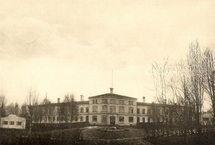 En svartvit, rektangulär bild visar en stor byggnad med flera våningar och skorstenar på taket. Marken framför byggnaden har en central, rund formation. Smala, kala träd omger byggnaden, med en mindre byggnad till vänster och en bar himmel ovanför.