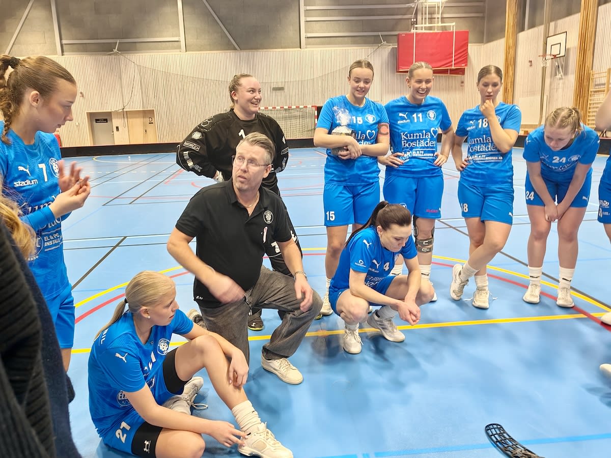 En grupp personer i idrottskläder, bestående av flera unga kvinnor i blå matchställ och en man i svart pikétröja, befinner sig på en idrottsplan inomhus. De flesta tittar mot mannen som sitter på huk, medan en kvinna sitter på planen och en innebandy