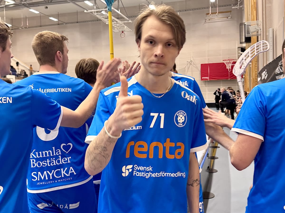 En grupp av män i blåa tröjor, troligtvis i en sporthall, med en man i förgrunden som ger en tummen upp. Logotyper och sponsornamn syns tydligt på tröjorna. I bakgrunden syns fler personer och inventarier som tyder på en idrottsmiljö.