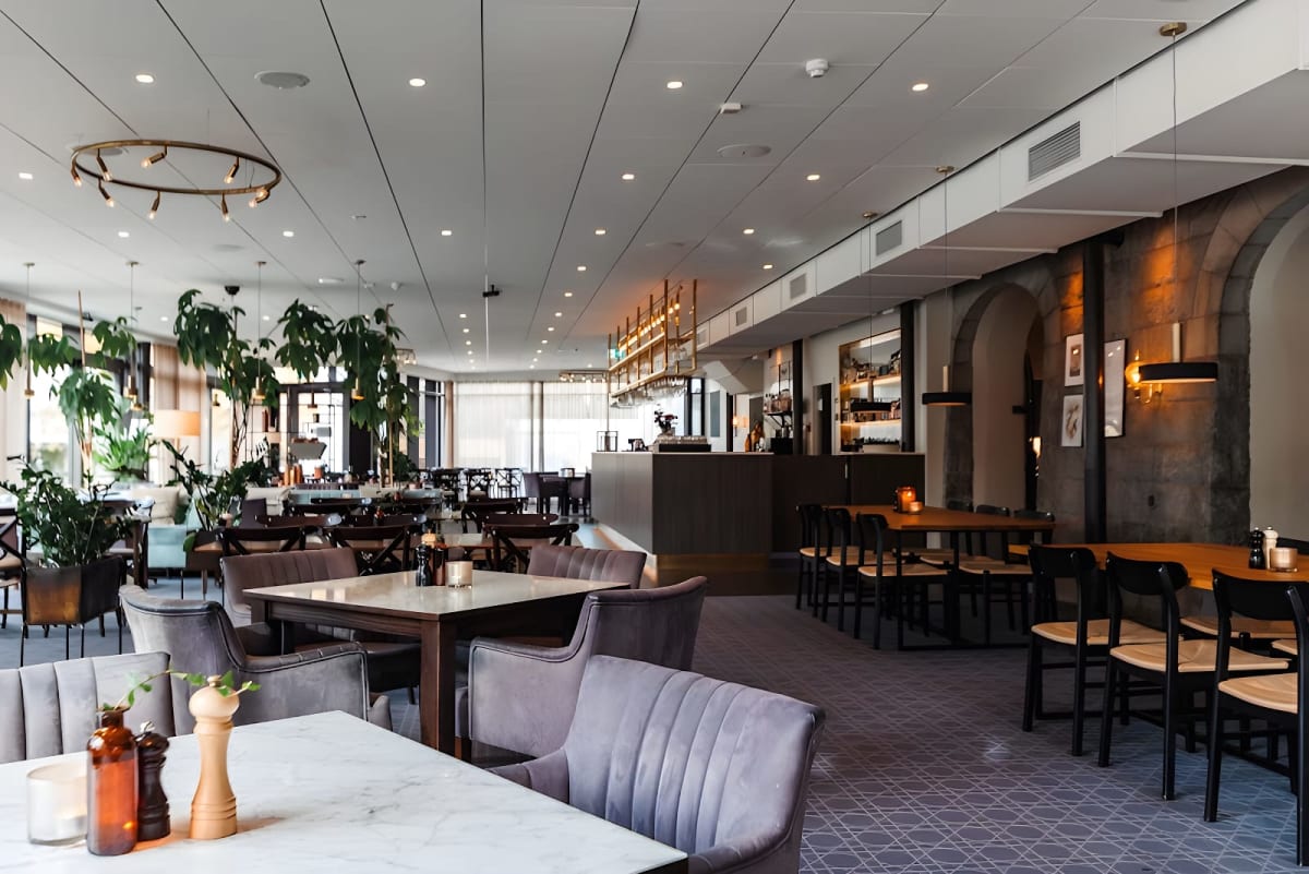 En stor, tom restauranglokal i ljusa färger, med flera bord och stolar. Till vänster syns växter och i bakgrunden en bar med hyllor och belysning. En grå mönstrad matta täcker golvet. Flera lampor hänger från taket.