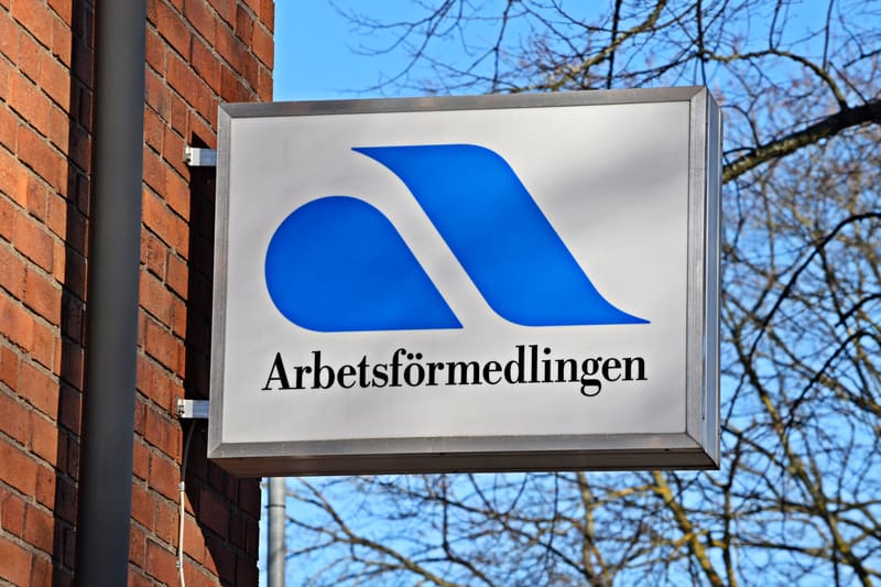 En rektangulär skylt med texten ”Arbetsförmedlingen” och en blå symbol är monterad på en röd tegelvägg. Bakom skylten syns trädgrenar utan löv och en blå himmel.