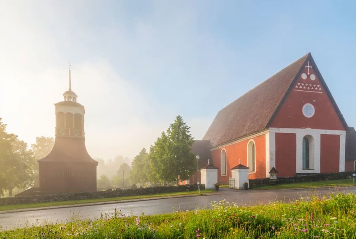 En röd kyrkobyggnad med ett rött tak och vita detaljer på fasaden, bredvid en fristående klockstapel i samma färg. En väg avgränsar stenmuren, med gräs och blommor i förgrunden. Morgondimma döljer delvis träd i bakgrunden.