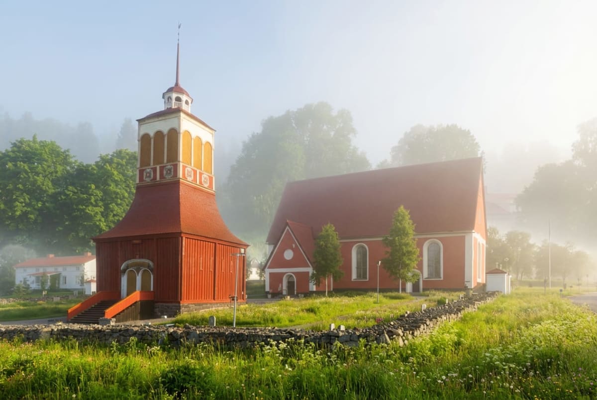 En röd kyrka med ett fristående klocktorn syns i bilden. Marken är grön med inslag av vildblommor. En stenmur avgränsar området. Dimma ligger över landskapet i bakgrunden, där träd och andra byggnader skymtar till. Solen skiner från höger.