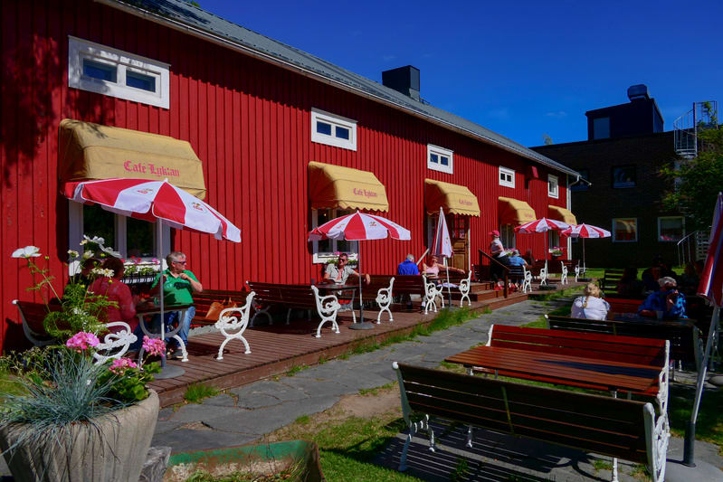 En röd träbyggnad med vita fönster och beigea markiser som det står "Cafe Lyktan" på, framför byggnaden finns ett utecafé med rödvita parasoller, mörka träbänkar och vita stolar. Flera personer sitter vid borden. En grön gräsmatta och en stensatt gån